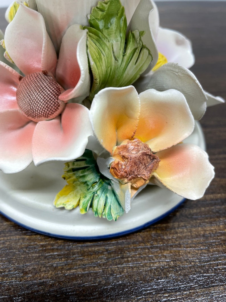 May include: A ceramic candle holder with a floral design. The holder features three pink, white, and orange flowers with green leaves. The base of the holder is white with a blue trim.