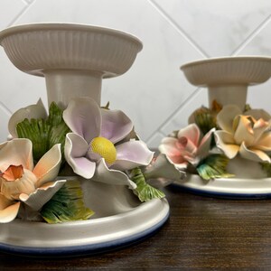 May include: Two white ceramic candle holders with floral accents. Each holder features a base with a ring of blue trim, a central column, and a top ring. The floral accents are made of porcelain and include pink, peach, and lavender flowers with green leaves.