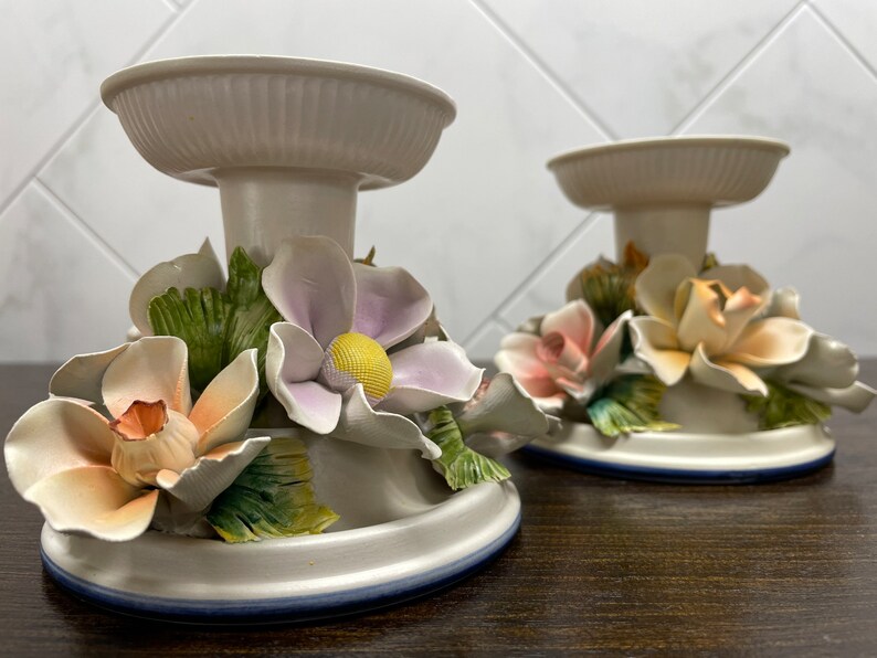 May include: Two white ceramic candle holders with floral accents. Each holder features a base with a ring of blue trim, a central column, and a top ring. The floral accents are made of porcelain and include pink, peach, and lavender flowers with green leaves.