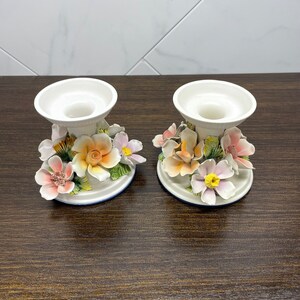 May include: A pair of white ceramic candle holders with floral designs. The holders feature pink, orange, and yellow flowers with green leaves.