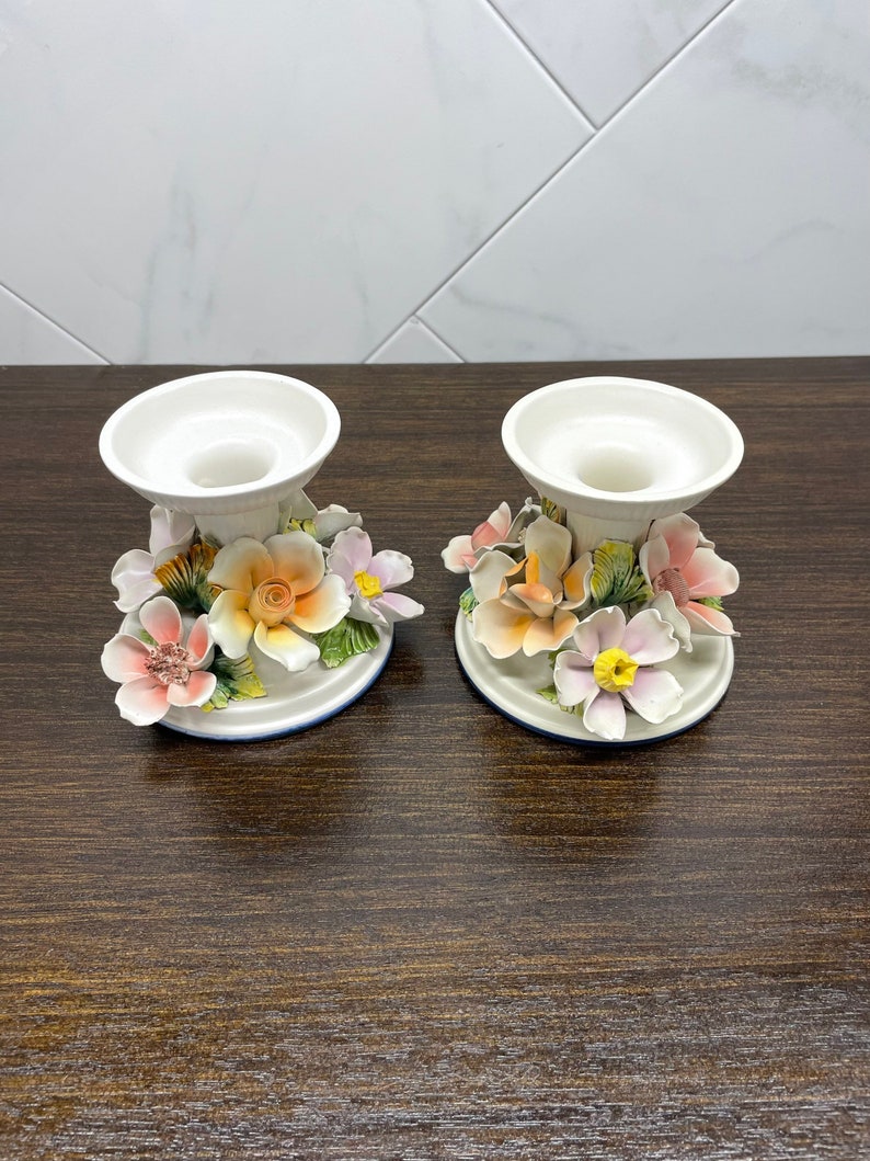 May include: A pair of white ceramic candle holders with floral designs. The holders feature pink, orange, and yellow flowers with green leaves.