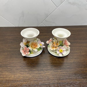 May include: Two white ceramic candle holders with floral designs. The holders feature pink, yellow, and orange flowers with green leaves.