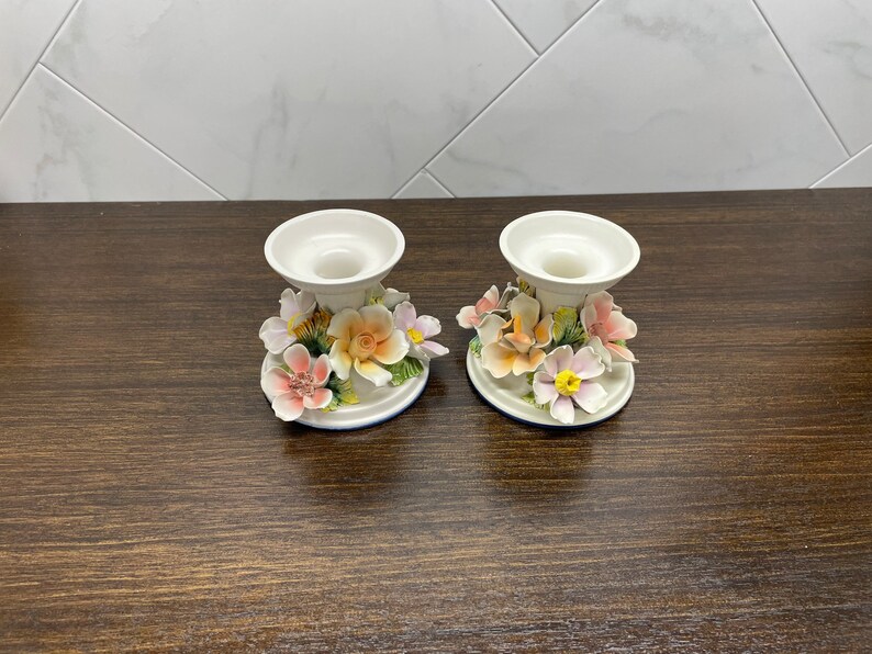 May include: Two white ceramic candle holders with floral designs. The holders feature pink, yellow, and orange flowers with green leaves.