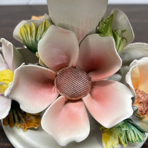 May include: A white ceramic candle holder with a floral design. The holder features a large pink flower with a brown center and smaller yellow and green flowers around it.