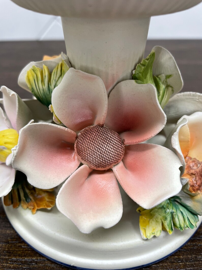 May include: A white ceramic candle holder with a floral design. The holder features a large pink flower with a brown center and smaller yellow and green flowers around it.