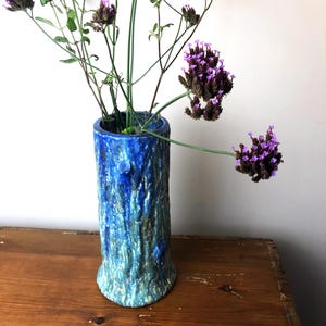 Peut inclure: Un vase en céramique bleu avec un extérieur texturé en forme d'écorce. Le vase contient des fleurs violettes avec des tiges vertes. Le vase est cylindrique avec une base légèrement évasée. Le vase mesure environ 20 cm de haut.