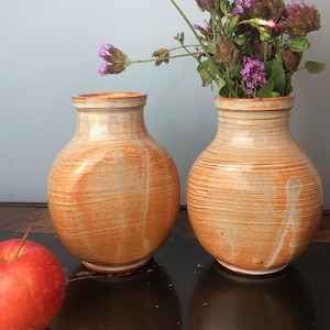 Peut inclure: Deux vases en céramique faits à la main avec une finition rustique et texturée dans des tons orange et crème. Un vase contient des fleurs violettes. Les vases ont un corps rond et un col étroit. Une pomme rouge repose sur la surface.