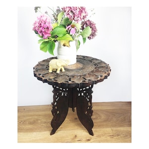 May include: A dark brown, intricately carved wooden side table with a round top. The table features a floral design around the edge and ornate legs. A white vase with pink flowers and a small elephant figurine sit on top.