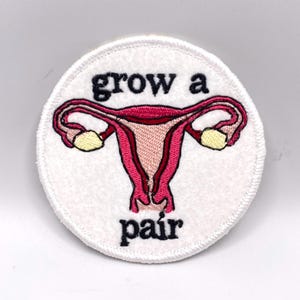 May include: A circular white embroidered patch featuring a pink and red anatomical uterus illustration in the center. The text "grow a" is stitched in black above the uterus, and the word "pair" is stitched in black below the uterus.