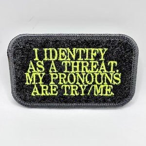 Puede incluir: Parche rectangular negro con el texto "I IDENTIFY AS A THREAT. MY PRONOUNS ARE TRY/ME" en hilo amarillo brillante. El parche tiene un borde negro y una superficie texturizada.