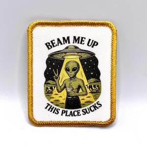 Könnte beinhalten: Rechteckiger Aufnäher mit goldenem Rand, der einen Außerirdischen mit einem Getränk zeigt, der ein Friedenszeichen gibt und unter einem UFO steht. Der Text lautet "BEAM ME UP" oben und "THIS PLACE SUCKS" unten.