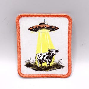 Könnte beinhalten: Quadratischer Aufnäher mit orangefarbenem Rand, der eine schwarz-weiße Kuh zeigt, die von einem gelben Strahl eines orange-schwarzen UFOs entführt wird. Der Hintergrund ist weiß.