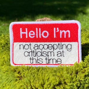 May include: A rectangular embroidered patch with a red top section featuring the white text Hello I'm and a white bottom section featuring the black text not accepting criticism at this time, all bordered by a thick white stitched edge.