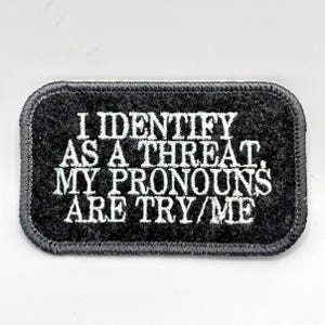 Puede incluir: Parche rectangular negro con texto bordado en blanco: "I IDENTIFY AS A THREAT. MY PRONOUNS ARE TRY/ME." El parche tiene un borde gris oscuro y una superficie texturizada. El parche está sobre un fondo blanco.
