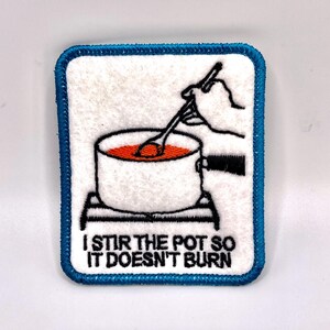 May include: Embroidered patch with a white background and a blue border. The patch features a black and white illustration of a pot with orange liquid being stirred by a hand. The text "I STIR THE POT SO IT DOESN'T BURN" is below the illustration.