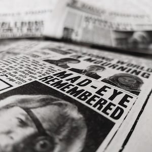 以下が含まれることがあります： 「MAD-EYE REMEMBERED」の見出しと「IN MOURNING」の文字が見える白黒の新聞ページ。顔とテキストのクローズアップショットが特徴で、ヴィンテージな雰囲気を醸し出しています。