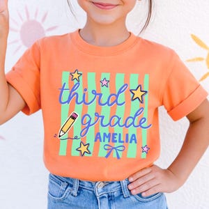 May include: An orange t-shirt with the words "third grade AMELIA" in blue script, a yellow pencil, and green stripes. The shirt is worn by a child wearing pink sunglasses and blue jeans. The background features a white wall with colorful sun designs.