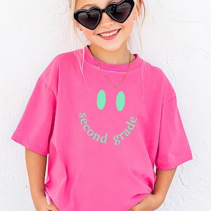 Può includere: Una ragazza indossa una maglietta rosa acceso con un disegno a faccina sorridente e la scritta "second grade". Indossa anche pantaloncini neri e occhiali da sole a forma di cuore. Il colore della maglietta è Crunchberry.