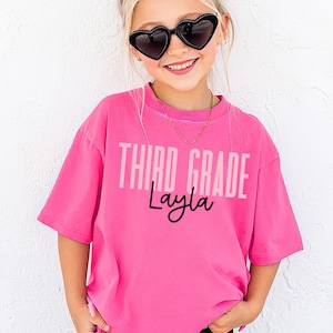 Peut inclure: Un t-shirt rose vif avec le texte "THIRD GRADE Layla" dans une police dégradée. Le t-shirt est porté par une jeune personne portant des lunettes de soleil en forme de cœur et un short noir. Le texte "Crunchberry" est en haut à droite.