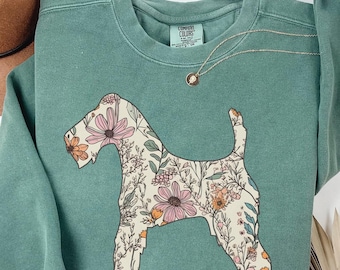 Puede incluir: Una sudadera verde salvia con una silueta floral de perro. El diseño del perro está lleno de flores y hojas rosas, naranjas y crema. También se ven un collar dorado y un sombrero marrón.