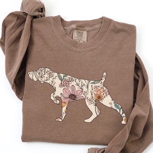 Op de afbeelding: Een bruin shirt met lange mouwen met een bloemenhondontwerp. De omtrek van de hond is gevuld met roze, oranje en groene bloemen. Het shirt is opgevouwen, met de mouwen geknoopt.