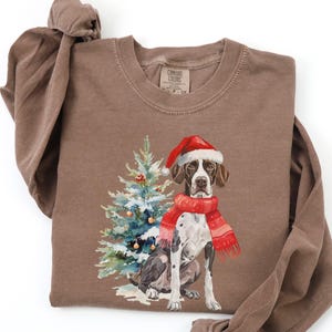 Könnte beinhalten: Ein braunes Langarmshirt mit einer Aquarellillustration eines Hundes mit Weihnachtsmütze und rotem Schal, zusammen mit einem geschmückten Weihnachtsbaum. Das Shirt ist aus weichem Material.