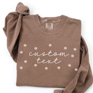 May include: A brown long-sleeve shirt with the words "custom text" in white script, surrounded by small white daisy graphics. The shirt is folded, with one sleeve tied in a knot.