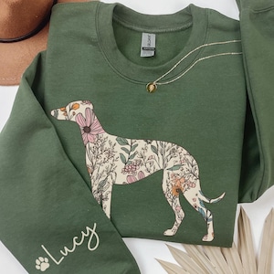 Peut inclure: Un sweat-shirt vert olive avec un motif de l&eacute;vrier floral. Le chien est rempli de fleurs roses, blanches et oranges. La manche porte le nom "Lucy" en cursive. Un collier en or et un chapeau marron sont &eacute;galement visibles.