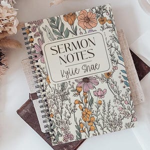 May include: A floral-patterned spiral-bound notebook labelled "Sermon Notes Kylie Shae" sits on a tray with a pen, a glass of liquid, and a bouquet of flowers. The notebook has a cream background with colourful flowers.