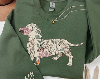 Könnte beinhalten: Grünes Sweatshirt mit floralem Dackel-Design. Der Hund ist mit rosa und weißen Blumen und Grün gefüllt. Auf dem Ärmel steht der Name "Lucy" in Schreibschrift. Eine goldene Halskette und ein brauner Hut sind ebenfalls zu sehen.