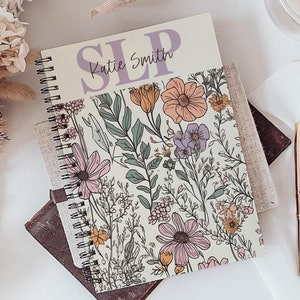 Puede incluir: Un cuaderno en espiral con estampado floral con el texto "SLP Katie Smith" se encuentra sobre una bandeja de madera con otros cuadernos. Un ramo de flores secas, un vaso de líquido y un bolígrafo dorado también están en la bandeja. Un difusor negro y una pequeña caja de regalo también son visibles.