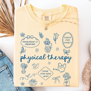 Puede incluir: Una sudadera de color mantequilla con la frase "physical therapy" en azul. La camisa presenta varias ilustraciones en azul, incluyendo flores, mariposas y las frases "Movement is Medicine" y "Every Step Counts."