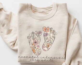 Può includere: Una felpa beige con un design floreale di due impronte di piedi e il testo "neonatal intensive care unit".