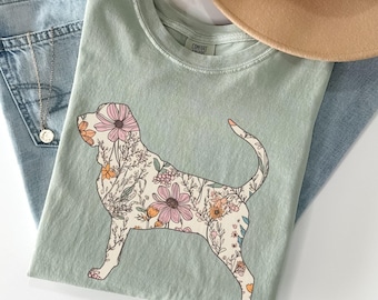 Könnte beinhalten: Ein salbeigrünes T-Shirt mit einem floralen Hunde-Design, kombiniert mit hellblauen Jeans und einem beigefarbenen Hut. Die Hunde-Grafik ist mit rosa, orangefarbenen und cremefarbenen Blumen gefüllt. Eine goldene Halskette und Trockenblumen vervollständigen das lässige, stilvolle Ensemble.