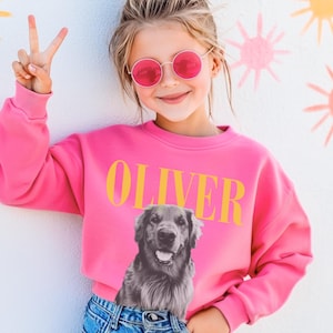 May include: A bright pink long-sleeve sweatshirt featuring the name OLIVER in bold yellow capital letters above a black and white photographic print of a golden retriever dog. The sweatshirt has a crew neckline and ribbed cuffs.