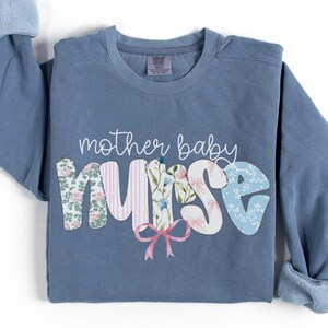 Könnte beinhalten: Ein blaues Sweatshirt mit dem Text "mother baby nurse" in Weiß. Das Wort "nurse" besteht aus bunten Stoff-Patches mit Blumenmustern.