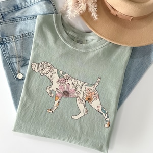 May include: A light green t-shirt with a floral print of a dog silhouette. The dog is a pointer breed with a long snout and long ears. The floral print is in shades of pink, orange, and yellow.