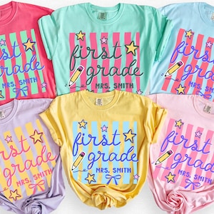 以下が含まれることがあります： Crunchberry、Island Reef、Chambray、Orchid、Butter、Blossomの6色の半袖Tシャツ。各シャツには、「first grade」と「MRS. SMITH」の文字、星、鉛筆、ストライプが描かれたグラフィックがプリントされています。