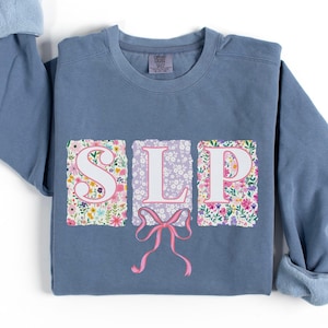 Puede incluir: Una sudadera azul claro con la palabra "SLP" escrita en letras blancas sobre un fondo con estampado floral. Un lazo rosa está centrado debajo de las letras.