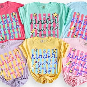 May include: Six colorful t-shirts in shades of pink, green, blue, purple, yellow, and pink. Each shirt features the words "kindergarten" with a pencil and star graphic. The shirts are labeled with color names.