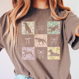 Puede incluir: Camiseta marrón con un diseño de cuadrícula que presenta siluetas de perros e ilustraciones de lazos. Las siluetas de perros están sobre fondos florales. La camiseta, de color neutro, es adecuada para el uso diario.
