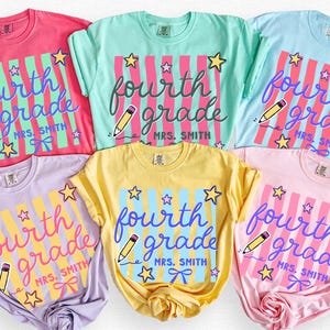 May include: Six short-sleeved t-shirts in various colors, including pink, blue, and yellow. Each shirt features the words "fourth grade" in a script font, along with a pencil and stars. The shirts are labeled with color names.