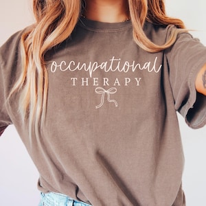 May include: A brown t-shirt with the words "occupational therapy" in a cursive font, with "THERAPY" in a block font. Below the text is a bow graphic. The shirt is a solid color and has short sleeves.