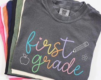 May include: A stack of various colored t-shirts. The top shirt is charcoal gray with the words "first grade" in rainbow script, along with a pencil, apple, and flower graphic. The shirts are made of a soft material.
