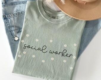 Può includere: Una maglietta verde chiaro con il testo "social worker" in corsivo nero. La maglietta è decorata con piccoli fiori di margherita bianchi.