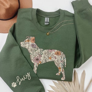May include: An olive green sweatshirt featuring a floral dog silhouette design. The dog is filled with colorful flowers. The sleeve has the name "Lucy" and a paw print. A gold necklace and a brown hat are also in the image.
