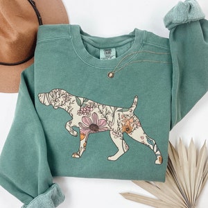 Puede incluir: Una sudadera verde con un estampado floral de una silueta de perro. La sudadera tiene cuello redondo y mangas largas. El estampado es en tonos de rosa, naranja y amarillo.