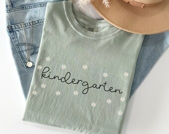 以下が含まれることがあります： 黒い筆記体で「kindergarten」と書かれた薄い緑色のTシャツ。Tシャツは小さな白いデイジーの花で飾られています。