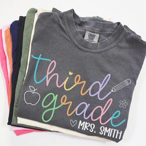 Op de afbeelding: Een stapel t-shirts in verschillende kleuren, met een donkergrijs shirt bovenop. Het bovenste shirt heeft de woorden "third grade" in kleurrijk schrift, samen met een appel, potlood, bloem en de naam "Mrs. Smith".
