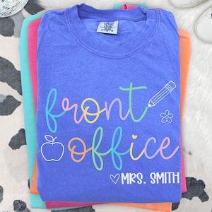 May include: A stack of colorful t-shirts, with a blue shirt on top. The blue shirt has the words "front office" in a rainbow script, along with a pencil, flower, and apple graphic. The name "Mrs. Smith" is below the words. The shirt is labeled "Flo Blue".